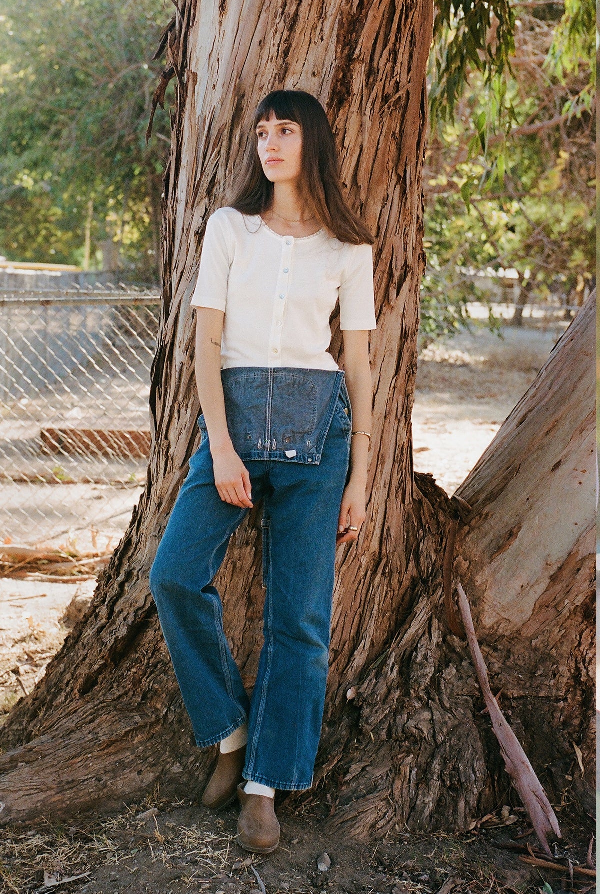 A woman with long hair leans against a tree trunk, wearing a Short Sleeve Organic Cardi from LA Relaxed. She is seen in blue jeans in a natural setting, with a metal fence visible in the background.