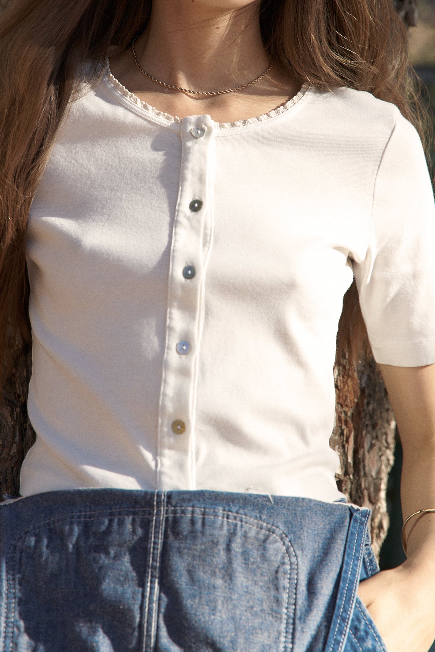 Wearing an LA Relaxed Short Sleeve Organic Cardi featuring shell buttons and a scalloped neckline, paired with a blue denim skirt, they stand by a tree, their long brown hair complementing a silver necklace.