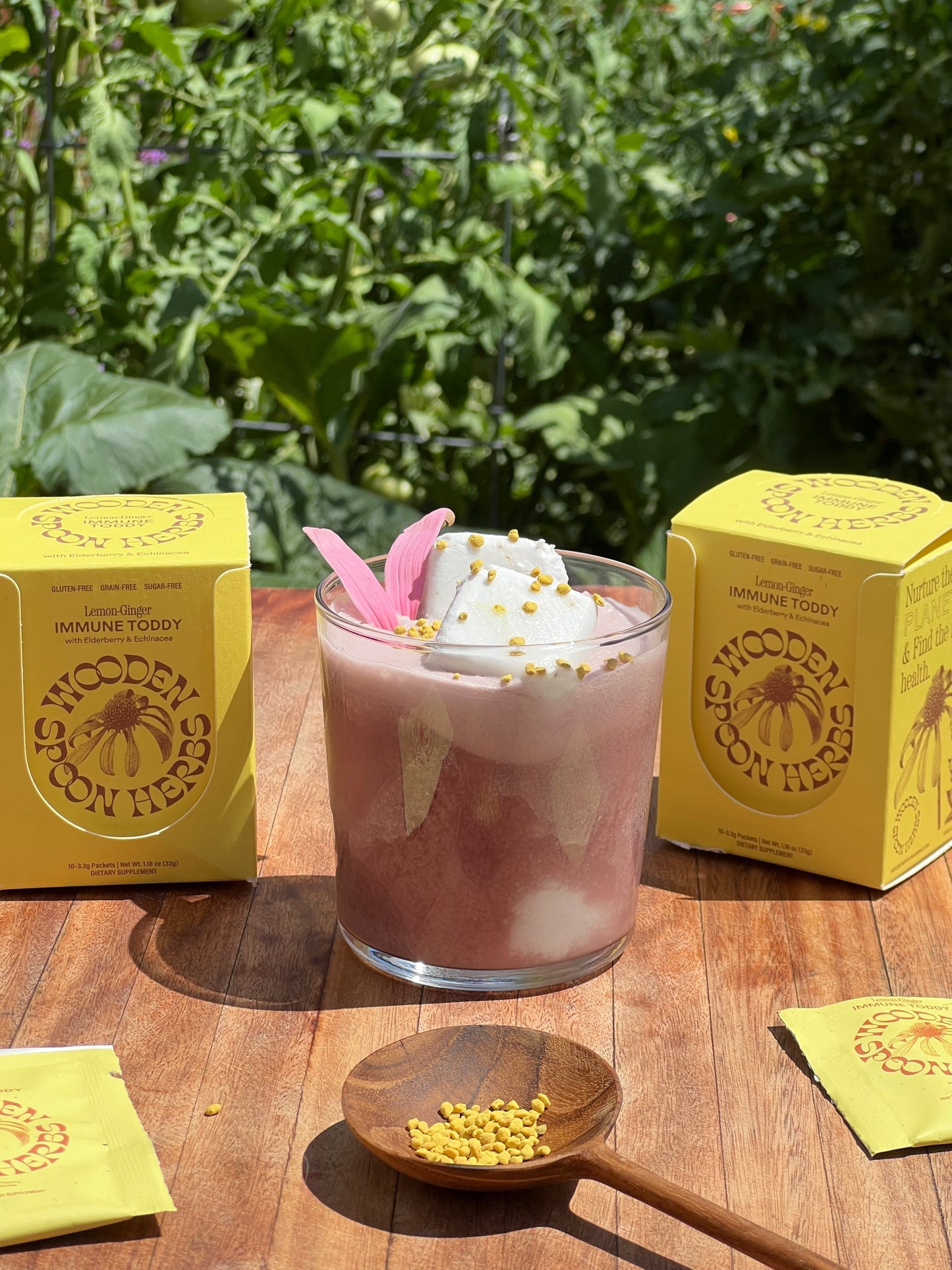 A refreshing Lemon-Ginger Immune Toddy Sachet drink topped with whipped foam and a pink flower sits on a wooden table. Yellow boxes labeled "WOODEN SPOON HERBS" surround the drink, offering immune support through their herbal remedies. A small wooden spoon with yellow granules is placed nearby. Lush green foliage is visible in the background.