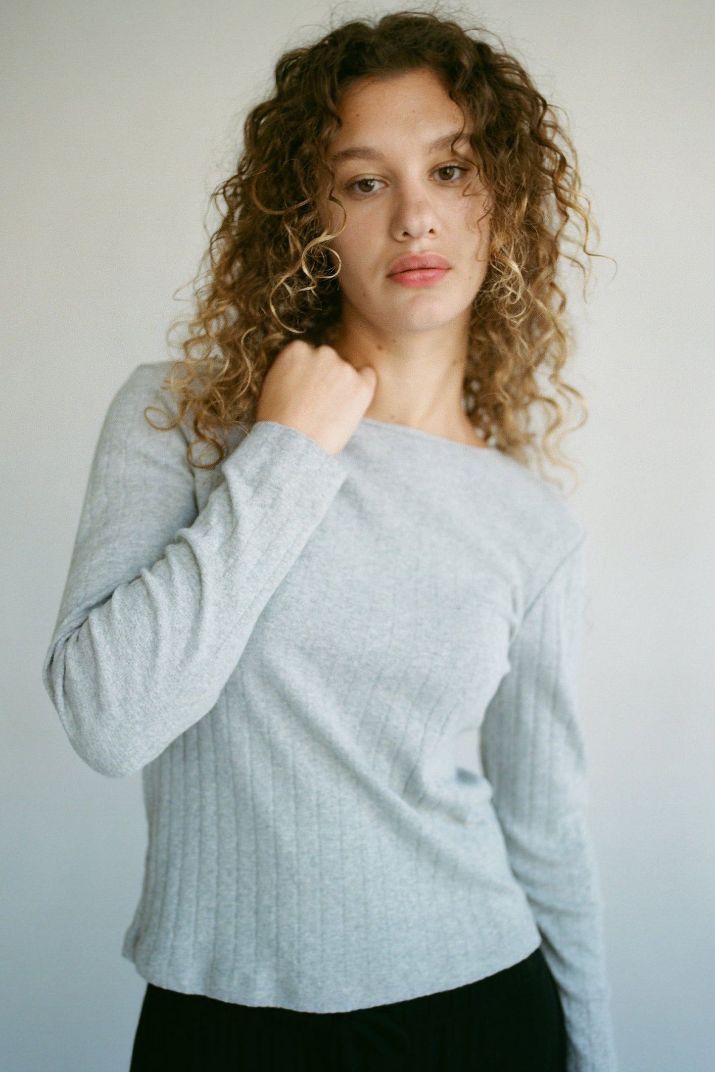 A woman with curly hair wears the LA RELAXED Organic Texture Long Sleeve in light grey, paired with black pants. Against a plain background, she gazes directly at the camera while her left hand gently touches her neck.