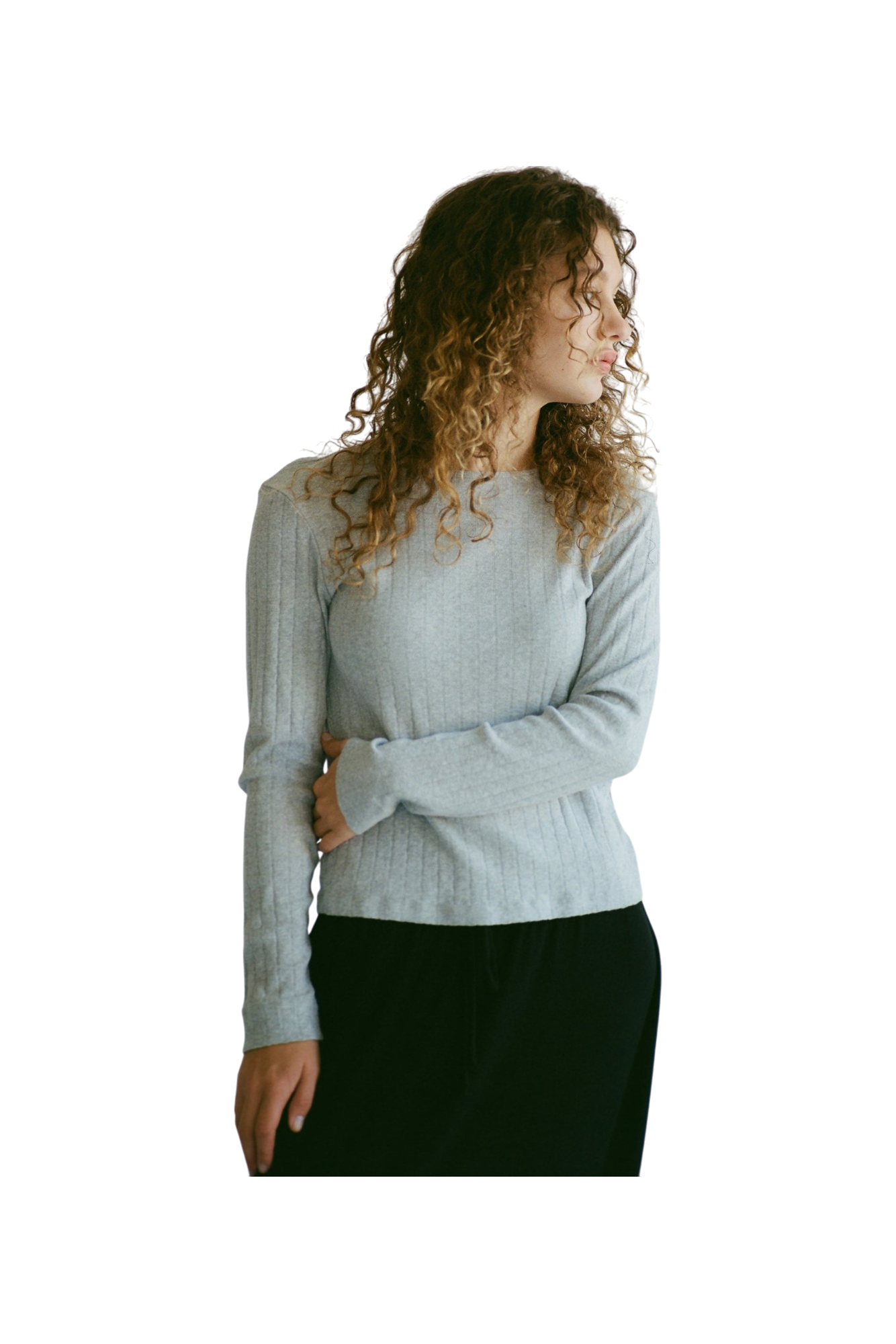 A person with curly hair wears the LA RELAXED Organic Texture Long Sleeve in light gray and dark pants. They stand with one arm crossed, gazing to the side against a plain background.
