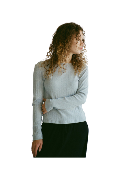 A person with curly hair wears the LA RELAXED Organic Texture Long Sleeve in light gray and dark pants. They stand with one arm crossed, gazing to the side against a plain background.