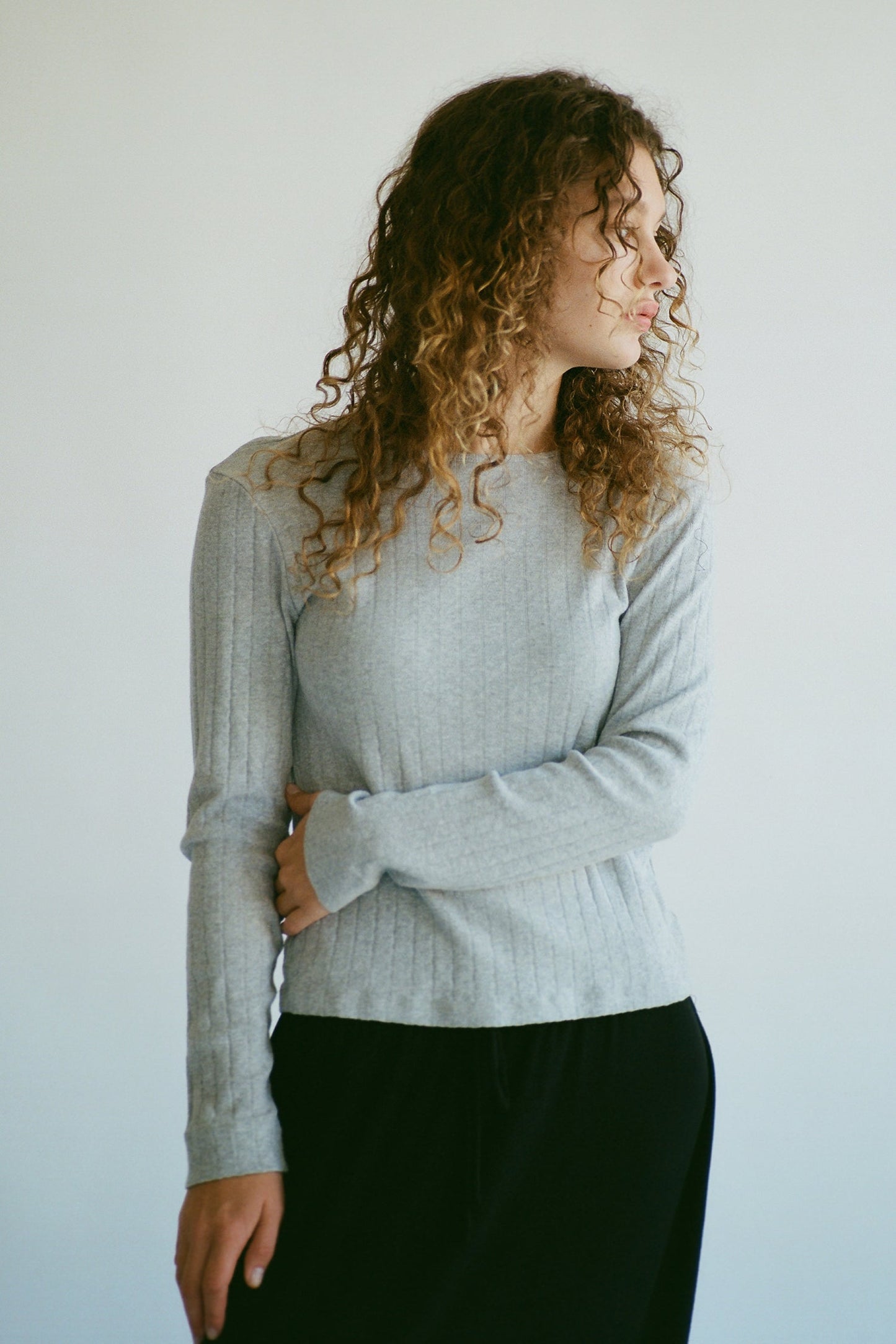 A person with curly hair stands against a light background, wearing the LA RELAXED Organic Texture Long Sleeve in gray and black pants. Their left arm is crossed over their body as they gaze thoughtfully to the side.