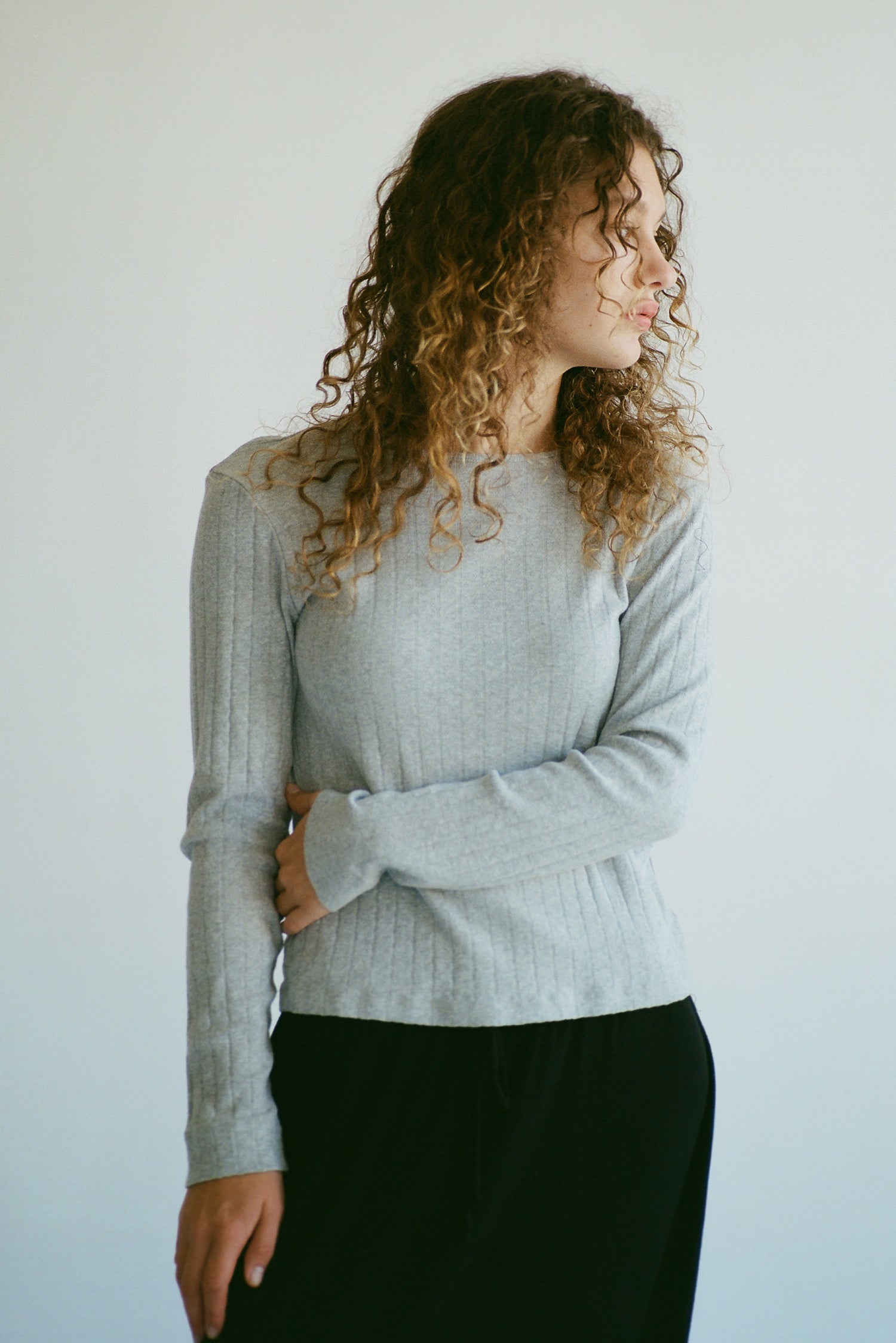 A person with curly hair stands against a light background, wearing the LA RELAXED Organic Texture Long Sleeve in gray and black pants. Their left arm is crossed over their body as they gaze thoughtfully to the side.