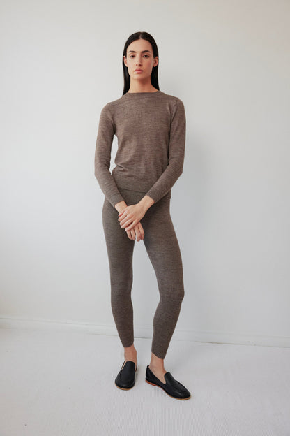 A person stands against a white background in a matching brown knit ensemble by Wol Hide, featuring the Layering Rib Crewneck in Heathered Brown with coordinating pants. Black loafers complete the look as they cross their arms comfortably.