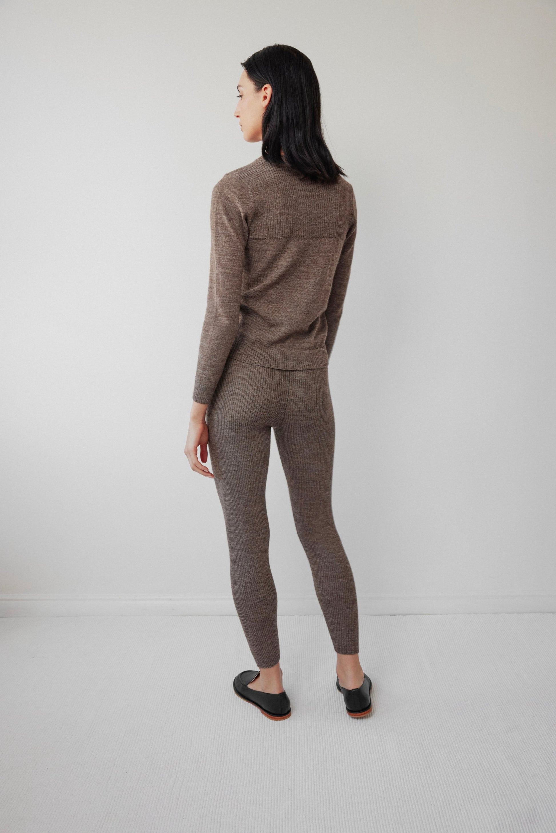 A woman stands before a blank wall in Wol Hide's Heathered Brown Layering Rib Crewneck and matching pants. Her shoulder-length straight black hair complements her ensemble, which includes black flat shoes on a light gray carpet.