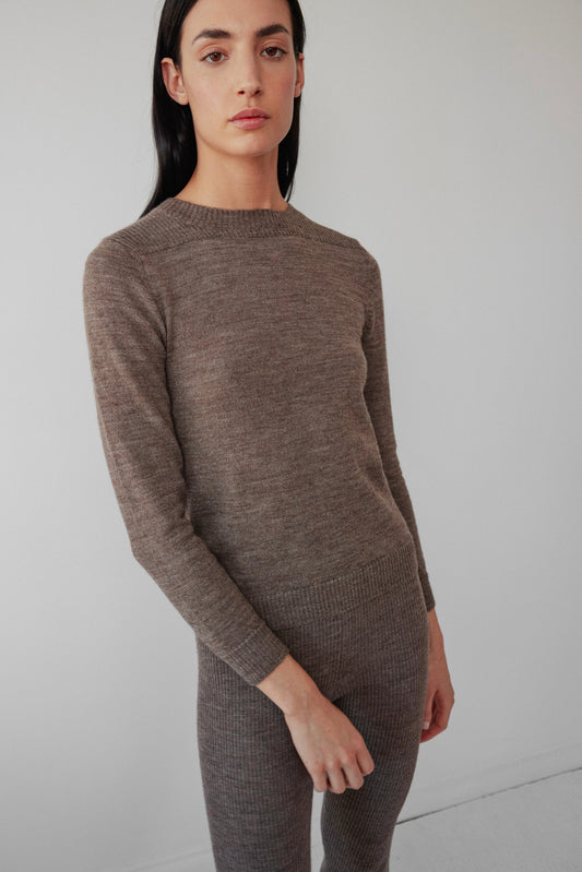 A person with long dark hair stands indoors, calmly facing the camera in Wol Hide’s Layering Rib Crewneck in Heathered Brown, which looks lightweight against a plain light-colored wall.