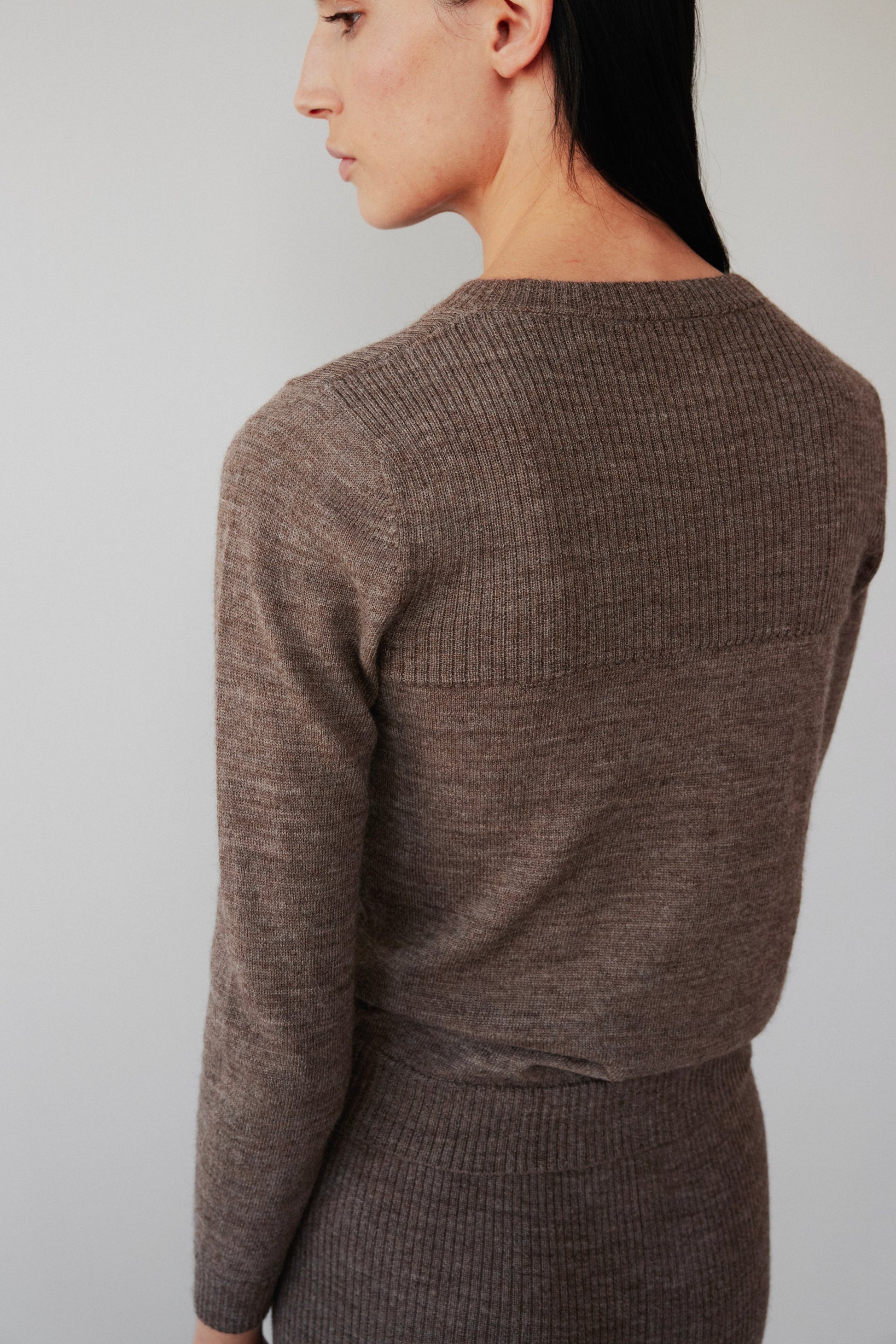 A person with long dark hair is turned to the side, wearing Wol Hide's Layering Rib Crewneck in Heathered Brown and a matching skirt. The plain, neutral background highlights the outfit's texture.