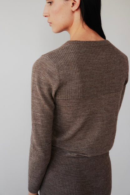 A person with long dark hair is turned to the side, wearing Wol Hide's Layering Rib Crewneck in Heathered Brown and a matching skirt. The plain, neutral background highlights the outfit's texture.