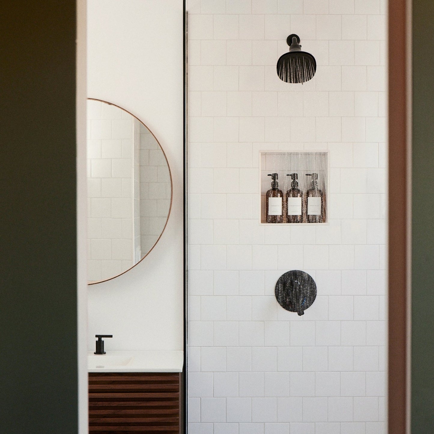 A minimalist bathroom featuring Canopy’s Filtered Showerhead, with clean lines, a round mirror over the sink, black fixtures, and natural tones. Three bottles on a built-in shelf enhance this serene, functional space.