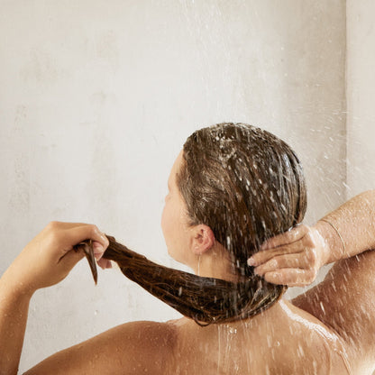 A person with wet brown hair stands under a Canopy Filtered Showerhead, holding their hair back and enjoying the soothing flow of filtered water, experiencing a moment of calm.