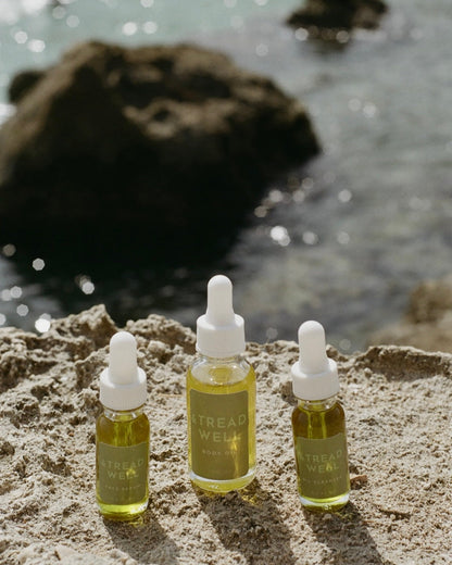 Three glass dropper bottles filled with yellow-green liquid labeled "& Tread Well Mini Essentials" rest on a sunlit sandy rock near the ocean, highlighting all-natural skincare against a backdrop of blurred water and rocks.