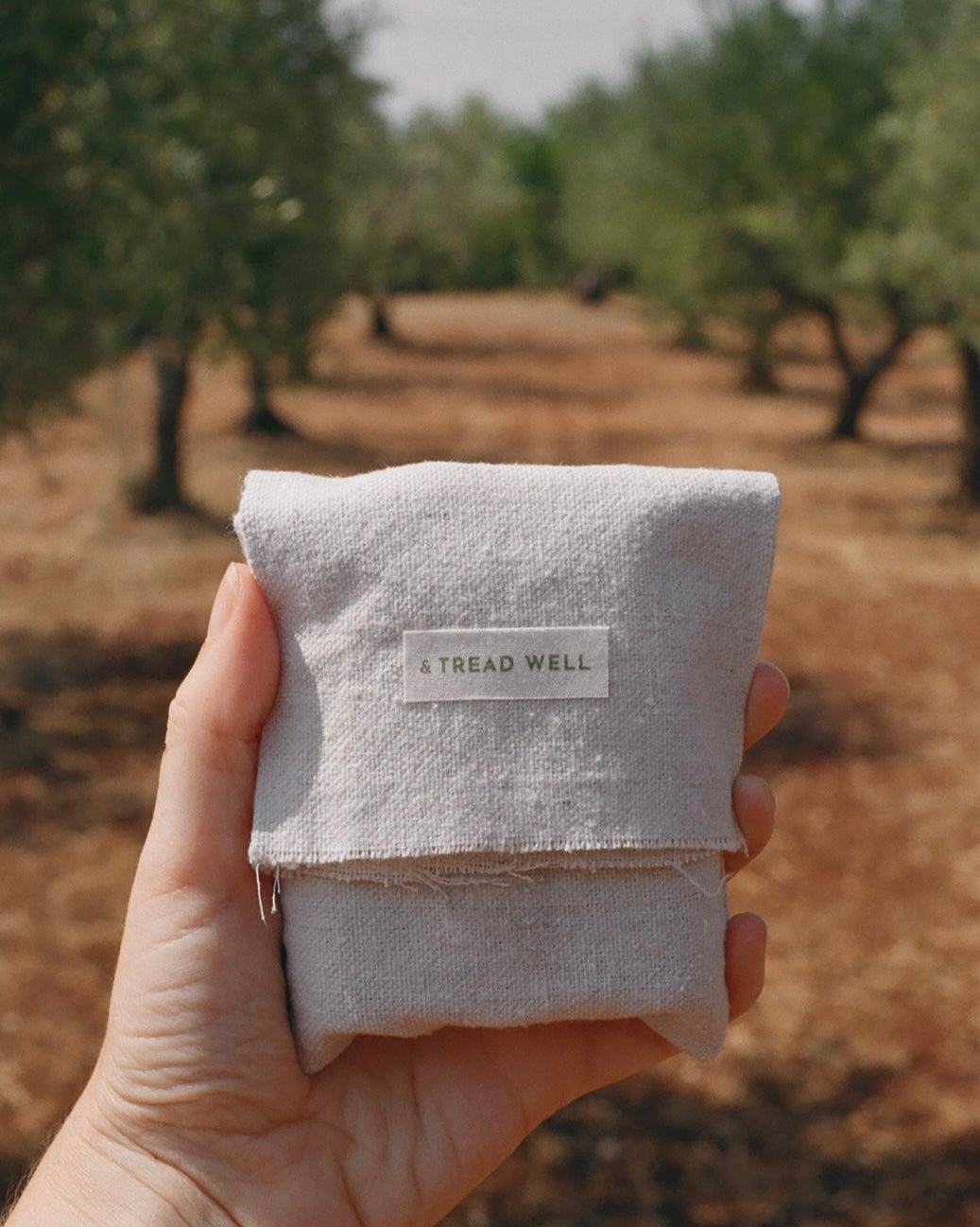A hand holds the Mini Essentials pouch by & Tread Well against an olive grove backdrop—the perfect setting for all-natural skincare.