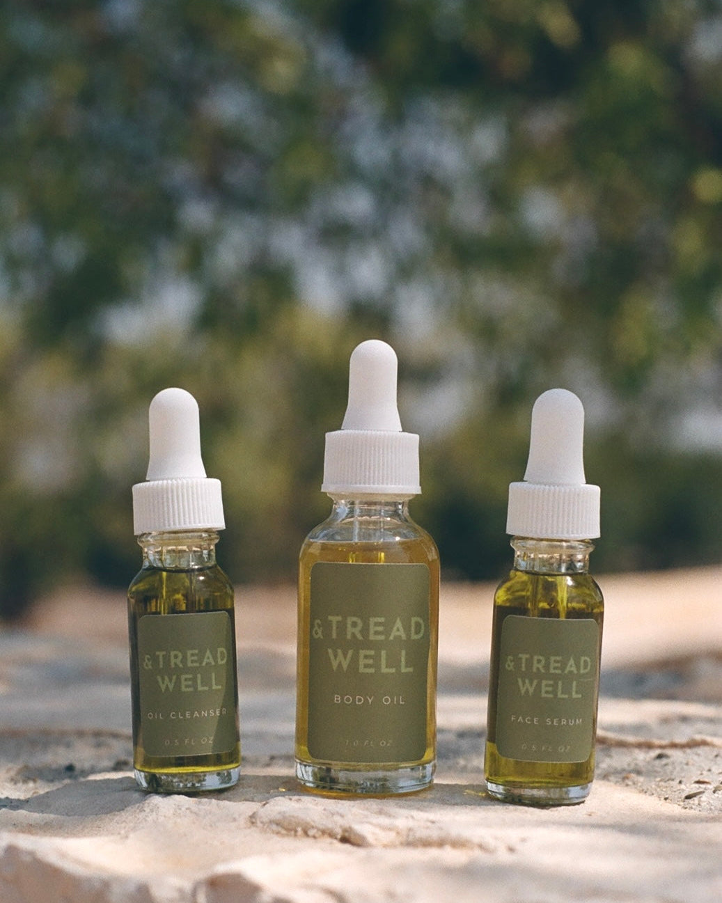 Three Mini Essentials by & Tread Well—all-natural skincare in glass dropper bottles—are arranged on a stone surface outdoors, with a blurred leafy background.