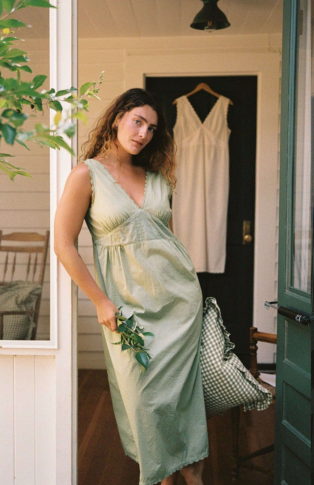 Mint - A woman with wavy hair calmly looks at the camera while wearing the Makinna Poplin Dress by LA RELAXED in light green, holding green leaves and a checkered cloth. Behind her, a white lace-trim dress hangs on the black door.