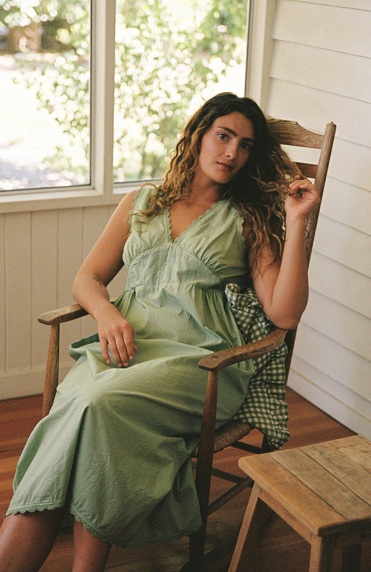Mint - A woman with long curly hair sits in a wooden rocking chair on a porch, wearing the Makinna Poplin Dress by LA RELAXED in light green. Sunlight streams through the window behind her as she gently holds a strand of her hair.