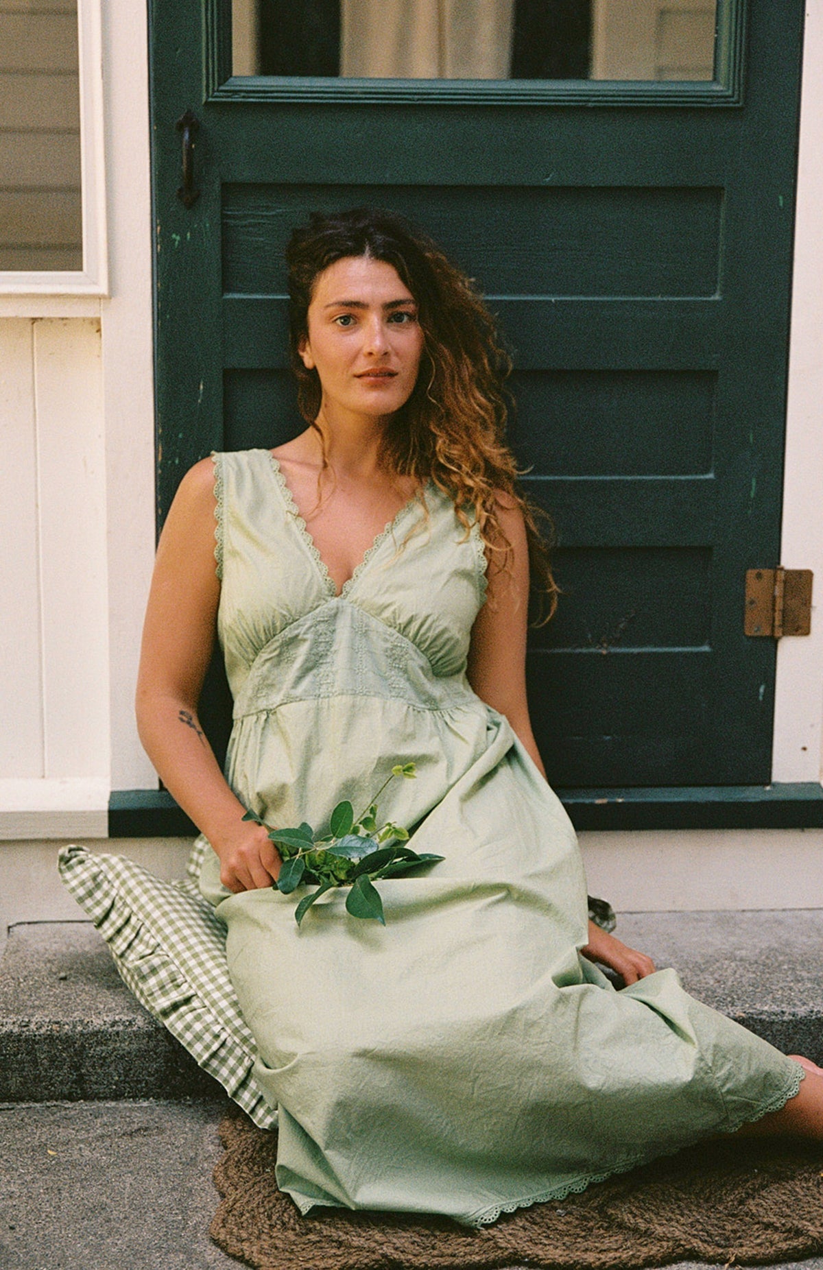Mint - A woman with wavy brown hair wearing the LA RELAXED Makinna Poplin Dress in light green sits on a doorstep holding green leaves, looking at the camera, with a green door and white wall behind her.