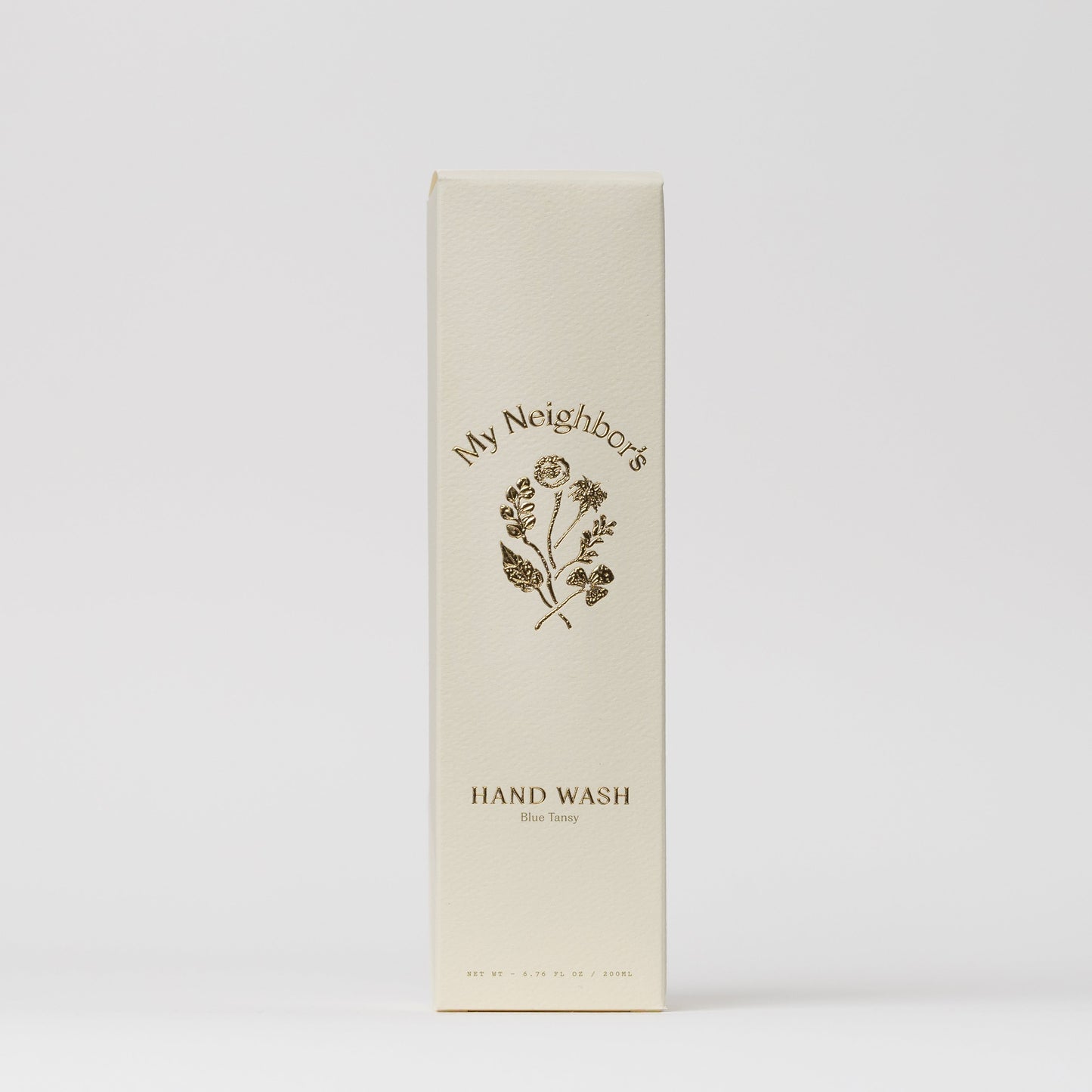 A cream-colored rectangular box labeled "My Neighbor’s Tallow" features minimalist gold text and a floral illustration, containing Blue Tansy Hand Wash. The packaging has a clean white background and an elegant design.