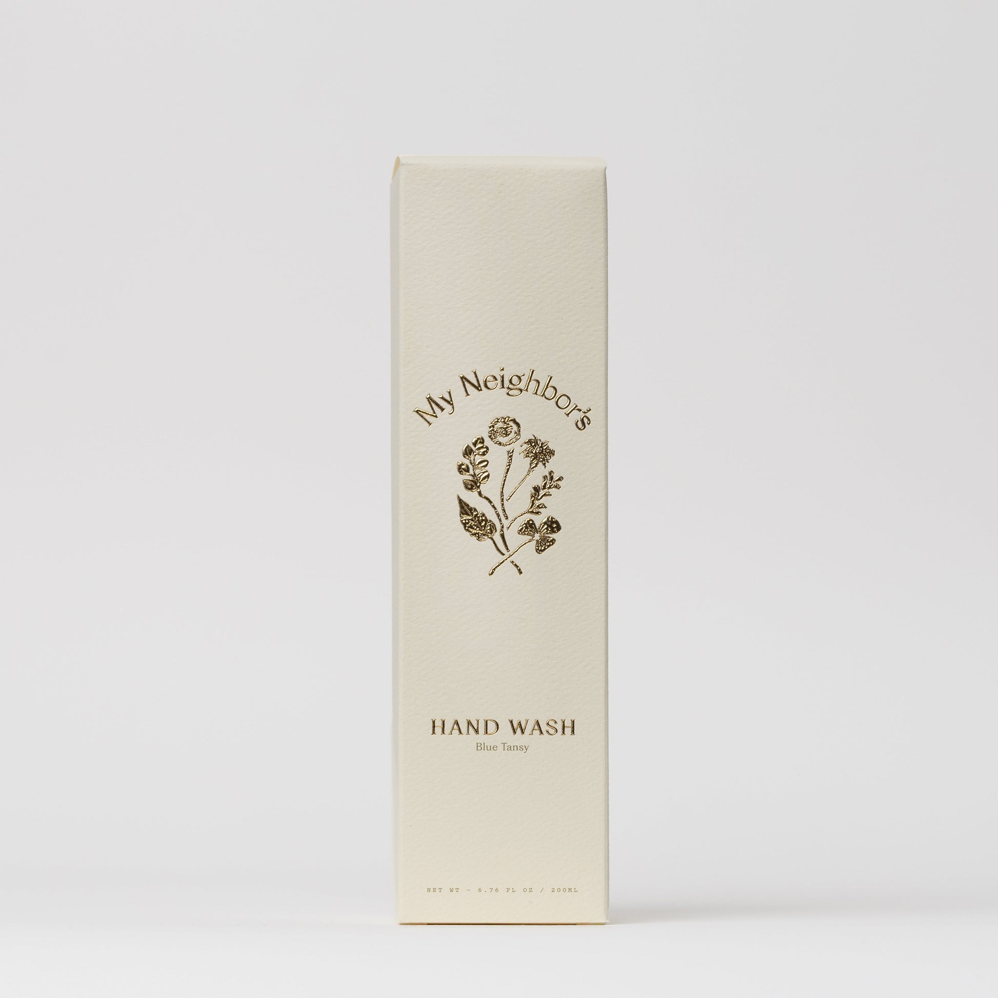 A cream-colored rectangular box labeled "My Neighbor’s Tallow" features minimalist gold text and a floral illustration, containing Blue Tansy Hand Wash. The packaging has a clean white background and an elegant design.