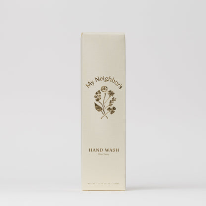 A cream-colored rectangular box labeled "My Neighbor’s Tallow" features minimalist gold text and a floral illustration, containing Blue Tansy Hand Wash. The packaging has a clean white background and an elegant design.
