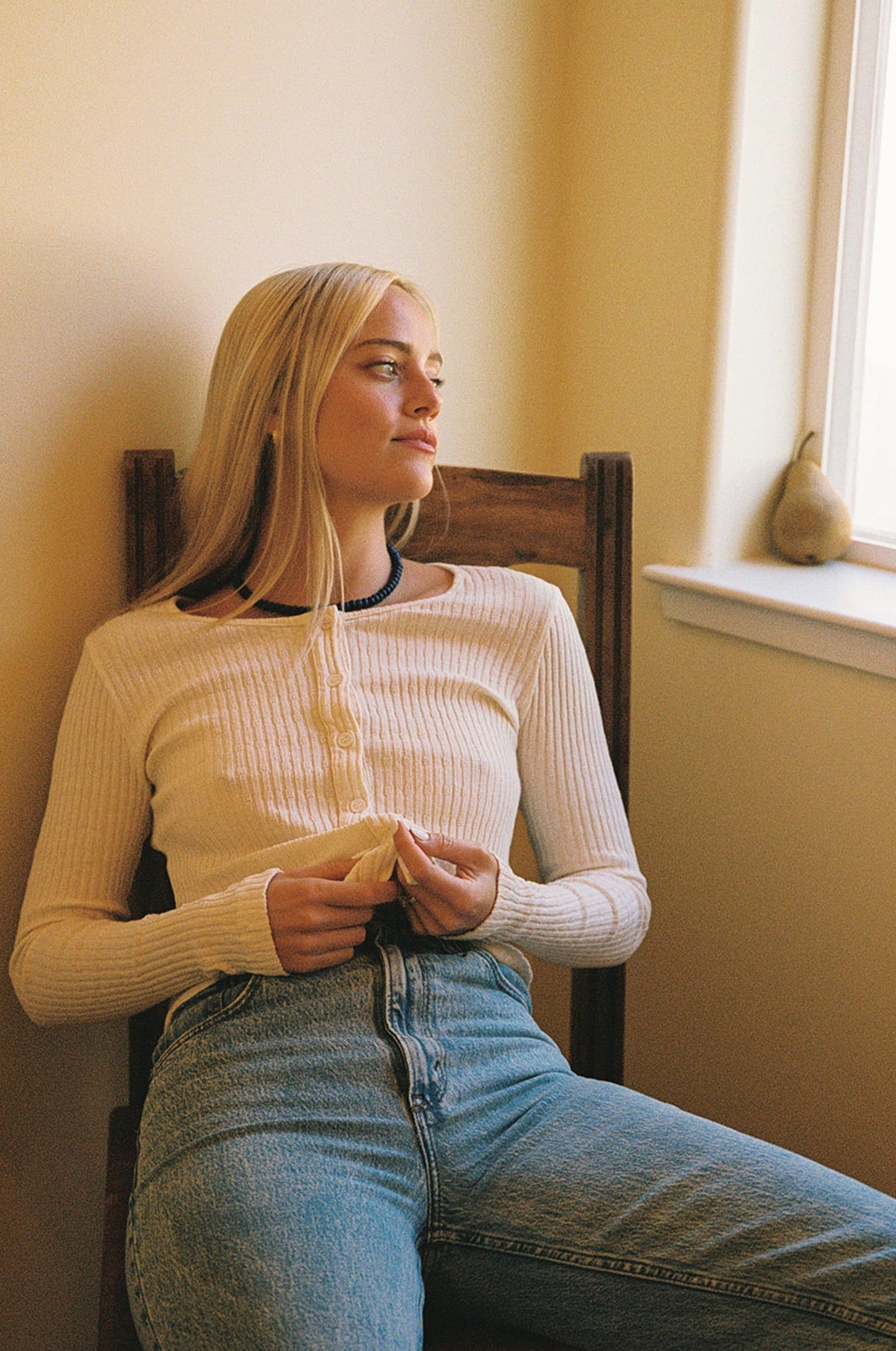 A woman with long blonde hair, wearing the LA RELAXED Pointelle Cardigan and blue jeans, sits on a wooden chair and looks thoughtfully out a window; a pear rests on the windowsill beside her.