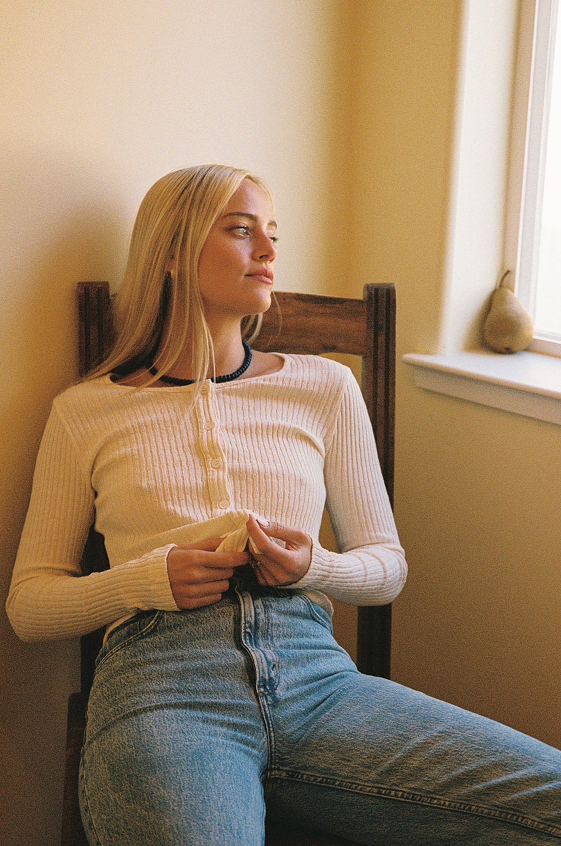 A woman with long blonde hair, wearing the LA RELAXED Pointelle Cardigan and blue jeans, sits on a wooden chair and looks thoughtfully out a window; a pear rests on the windowsill beside her.