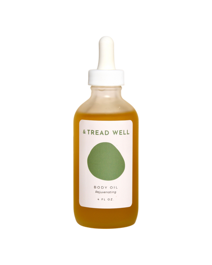 A clear glass dropper bottle of & Tread Well Body Oil, filled with golden oil and featuring a white and green label, designed to nourish skin, is shown against a black background.