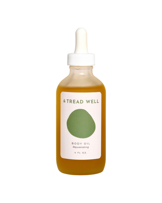 A clear glass dropper bottle of & Tread Well Body Oil, filled with golden oil and featuring a white and green label, designed to nourish skin, is shown against a black background.