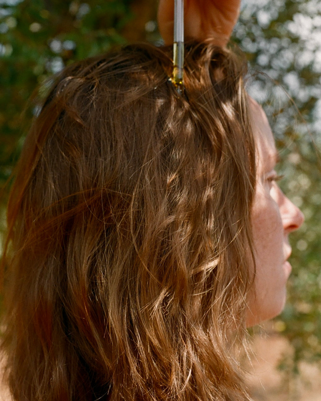A person with wavy brown hair is shown outdoors while & Tread Well Hair Oil is applied to their scalp with a dropper, nourishing strands for hair growth. Sunlight filters through greenery in the background.