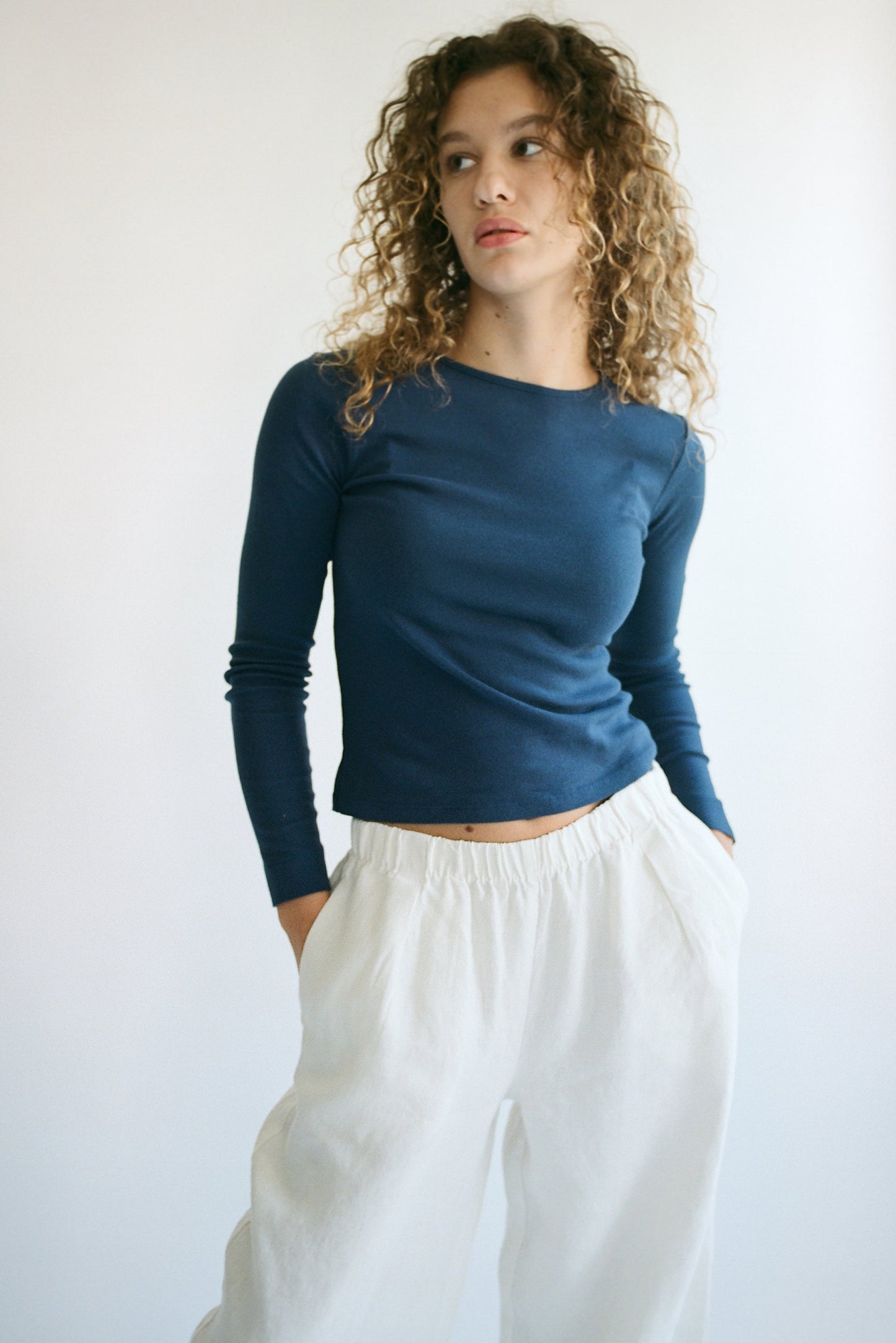 A person with curly hair is wearing an LA RELAXED Bebe Organic Long Sleeve navy top and white pants. They stand against a plain light background with their hands in their pockets, looking slightly to the side.