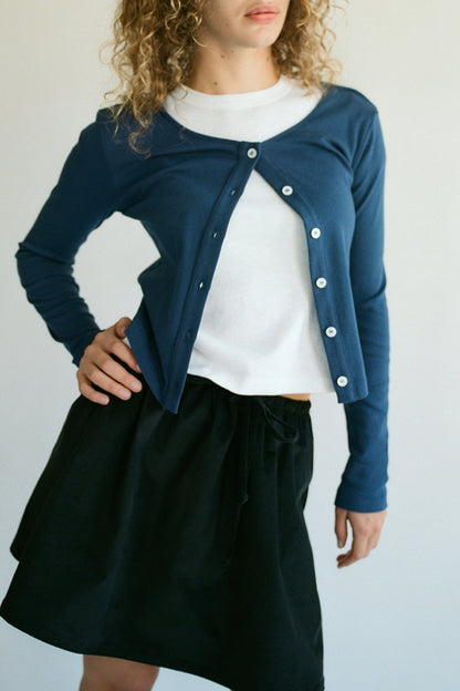 Navy 
Channeling a 90's Closet vibe, a model stands with hand on hip wearing LA Relaxed's blue Organic Rib Cardigan over a white top and dark skirt. The cardigan is stylishly half-unbuttoned, showcasing its organic cotton fabric against a plain light backdrop. Curly hair completes the look.