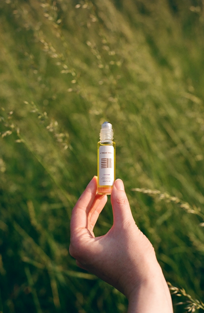A hand holds a bottle of & Tread Well Natural Fragrance - Nectar with a rollerball applicator, against blurred green grass in sunlight.