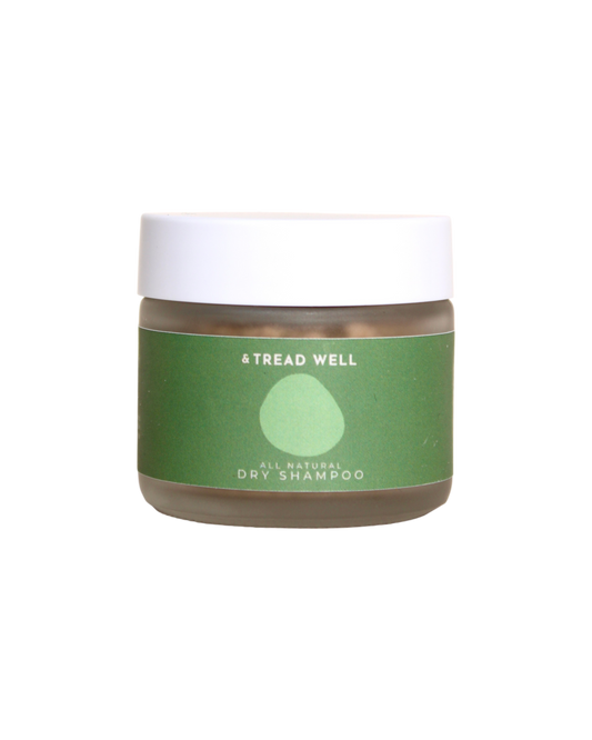 A small clear jar with a white lid and a green label reading "& Tread Well Dry Shampoo." This non-aerosol, plant-based hair powder from & Tread Well features a simple leaf or droplet graphic on its packaging.