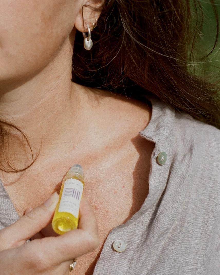 A person in a light gray button-up shirt applies & Tread Well’s Natural Fragrance - Nostalgia oil with a rollerball to their collarbone, showing only their lower face, neck, and hand.