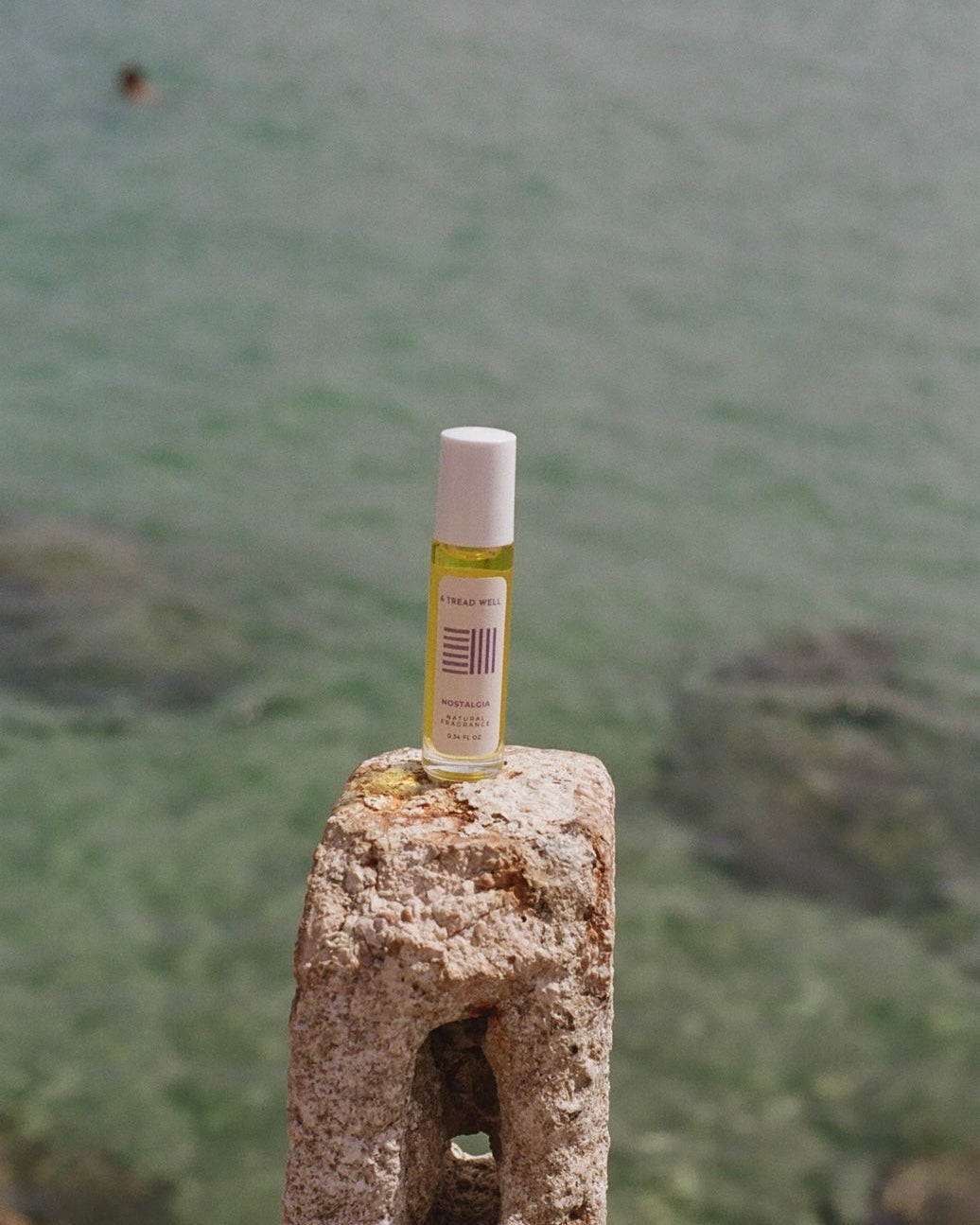 A small bottle of & Tread Well's Natural Fragrance - Nostalgia sits on a weathered stone post by the sea, its pure scent harmonizing with the clear water and rocky shoreline.