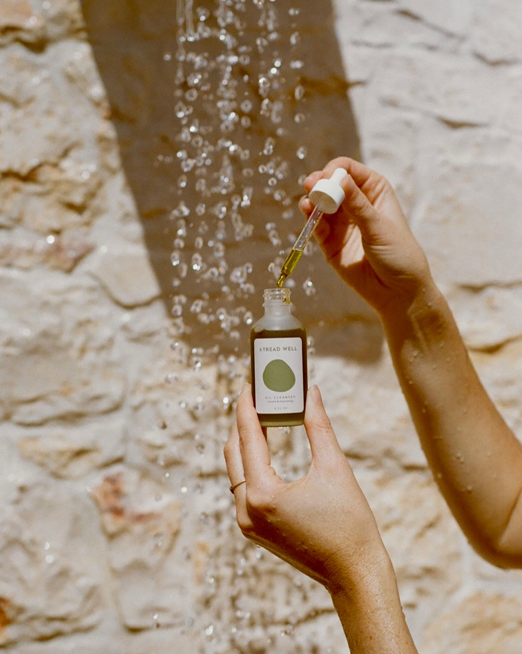 A person holds a dropper above an Oil Cleanser bottle labeled "& Tread Well," with water droplets in the background against a light stone wall.