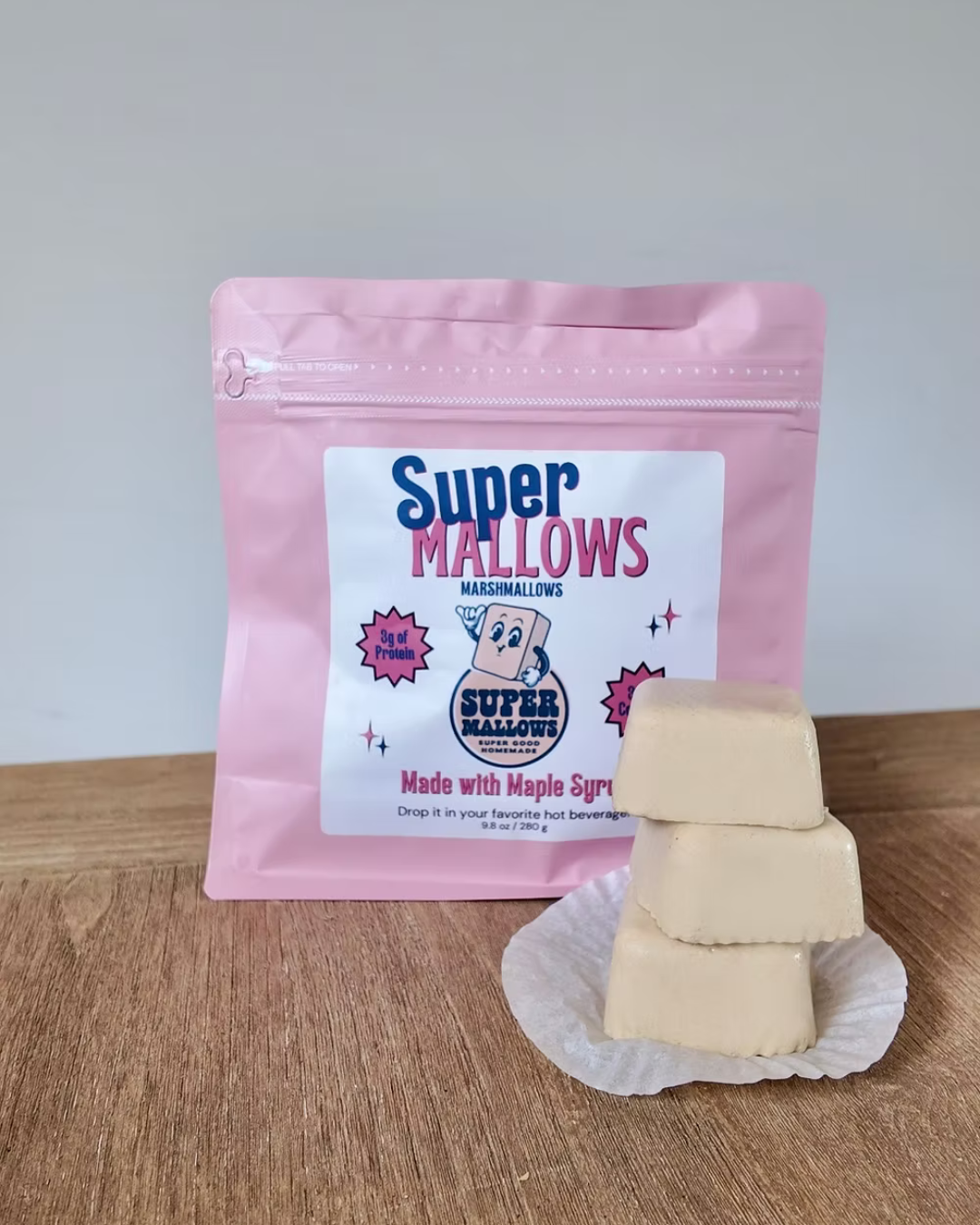 A pink resealable bag of Super Mallows Original Super Mallow 8-pack sits behind three stacked collagen marshmallows on a white wrapper, all set on a wooden surface with a neutral background.