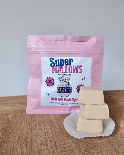 A pink resealable bag of Super Mallows Original Super Mallow 8-pack sits behind three stacked collagen marshmallows on a white wrapper, all set on a wooden surface with a neutral background.