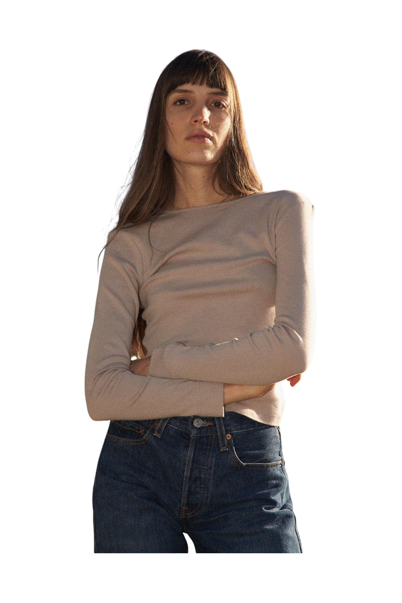 A woman with long hair and bangs, wearing a 90's inspired LA RELAXED Bebe Organic Long Sleeve in beige and blue jeans, stands with arms crossed against a black background.