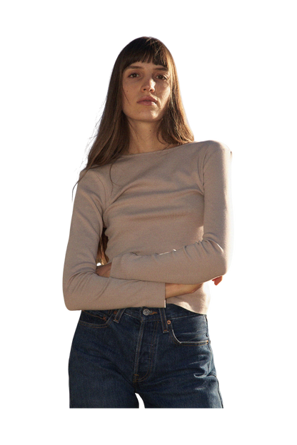 A woman with long hair and bangs, wearing a 90's inspired LA RELAXED Bebe Organic Long Sleeve in beige and blue jeans, stands with arms crossed against a black background.