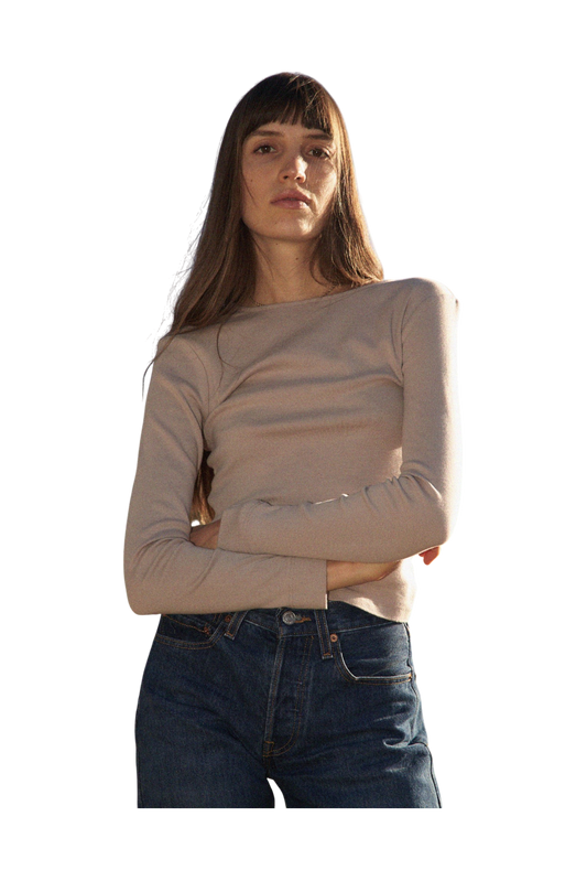 A woman with long hair and bangs, wearing a 90's inspired LA RELAXED Bebe Organic Long Sleeve in beige and blue jeans, stands with arms crossed against a black background.