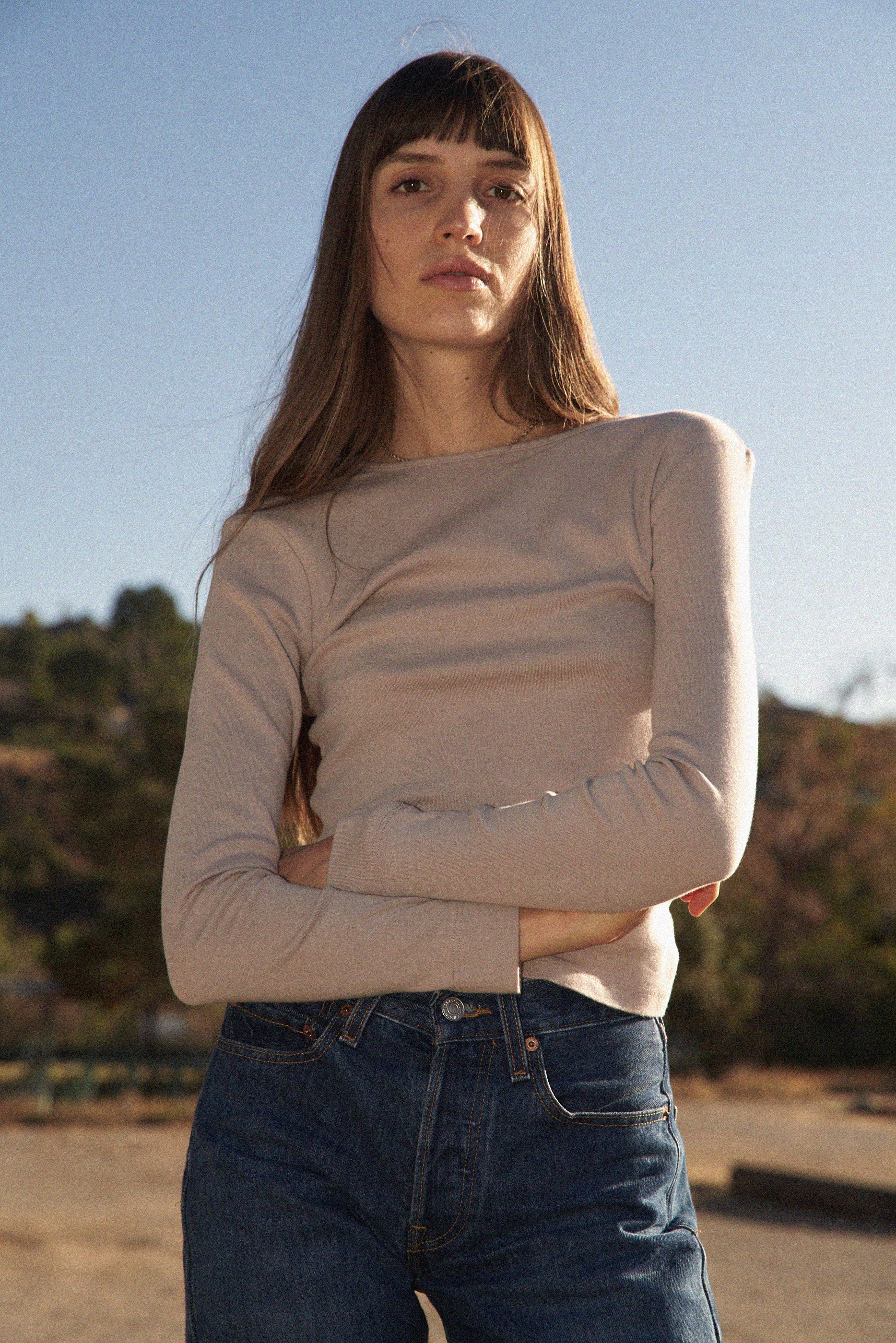 A person with long brown hair stands outdoors, confidently gazing at the camera amidst trees and a clear sky. They wear a Bebe Organic Long Sleeve by LA RELAXED, paired with blue jeans, their arms crossed.