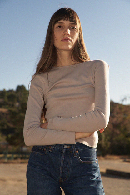 A person with long brown hair stands outdoors, confidently gazing at the camera amidst trees and a clear sky. They wear a Bebe Organic Long Sleeve by LA RELAXED, paired with blue jeans, their arms crossed.