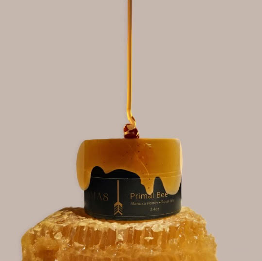 A jar of Wild Mamas' Wildly Nourishing Bison Tallow Balm – Primal Bee (Manuka & Royal Jelly) sits on honeycomb, honey streaming down its side and from above, evoking nourishing tallow skincare against a plain, neutral background.