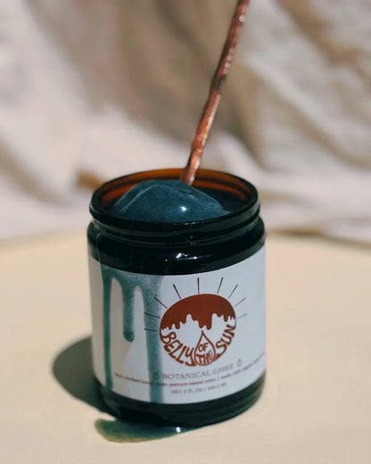 A brown glass jar labeled "Cookma Mama Bundle" sits on a light surface, filled with dark green ghee. A metal spoon is dipped in, ideal for making nourishing postpartum meals that support recovery and wellness.