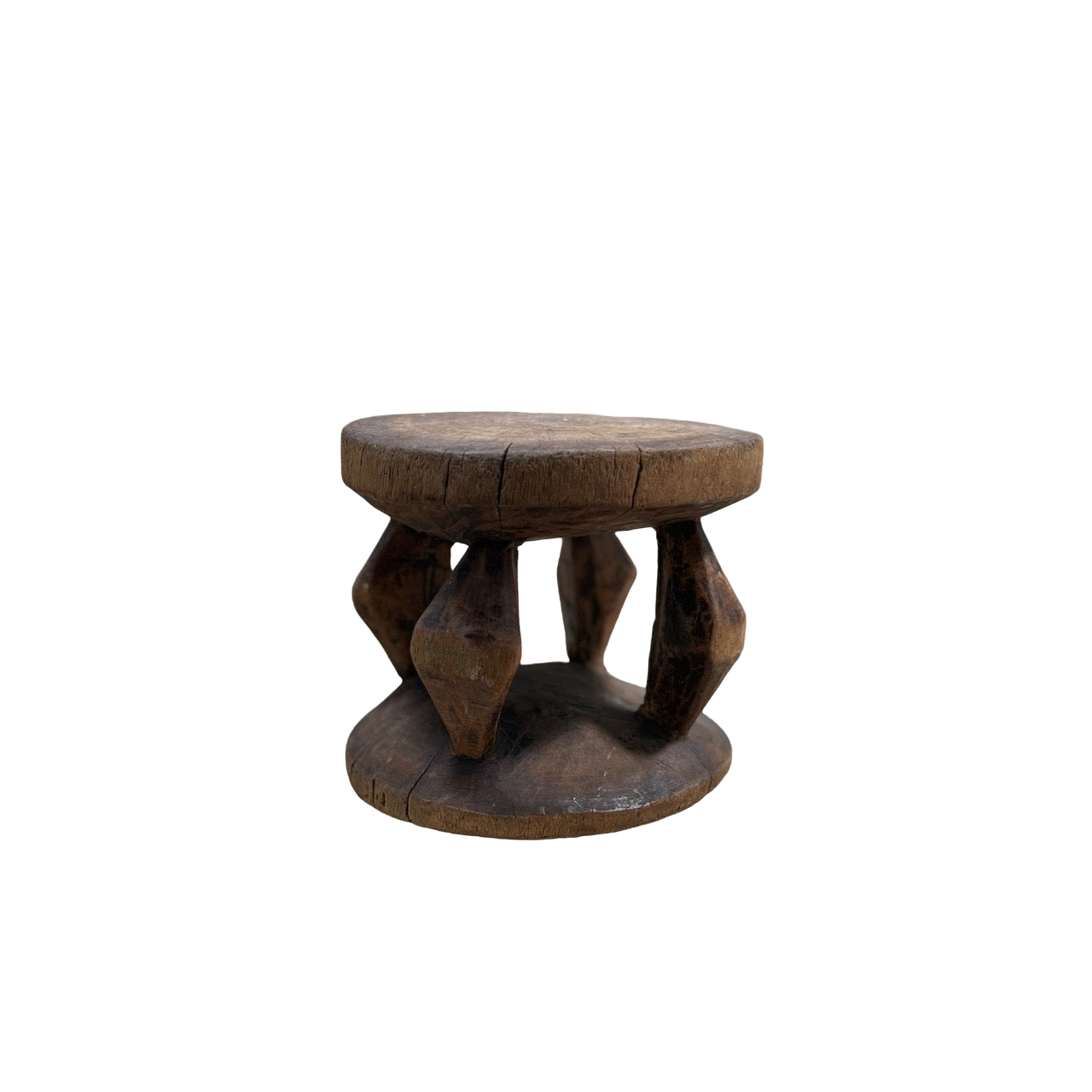 The Vintage African Wood Mini Stool by Shoppe Maison is a small, rustic wooden stool featuring a round top and bottom. It's connected by four intricately carved legs that display geometric shapes in African-inspired designs. The stool's weathered appearance, with visible cracks and wear, enhances its vintage look against the black background.