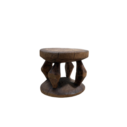 The Vintage African Wood Mini Stool by Shoppe Maison is a small, rustic wooden stool featuring a round top and bottom. It's connected by four intricately carved legs that display geometric shapes in African-inspired designs. The stool's weathered appearance, with visible cracks and wear, enhances its vintage look against the black background.