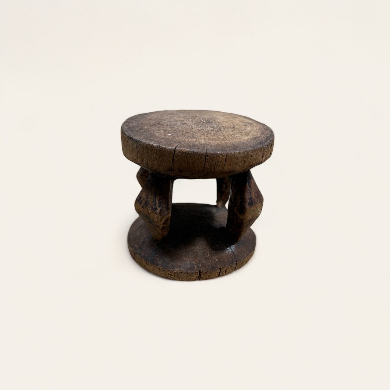 The Vintage African Wood Mini Stool by Shoppe Maison features a small, round wooden design with a rustic appearance. Its flat top and bottom are connected by three thick support posts carved from African wood. The stool showcases visible cracks and a weathered surface, adding to its aged charm. This vintage piece is displayed against a plain background.