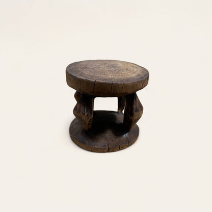 The Vintage African Wood Mini Stool by Shoppe Maison features a small, round wooden design with a rustic appearance. Its flat top and bottom are connected by three thick support posts carved from African wood. The stool showcases visible cracks and a weathered surface, adding to its aged charm. This vintage piece is displayed against a plain background.
