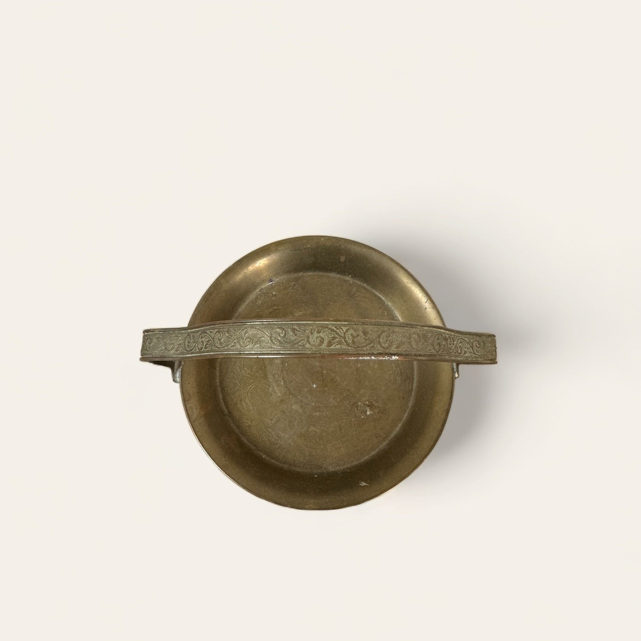 An overhead view of the Mini Brass Tray by Shoppe Maison reveals a vintage brass piece with a single horizontal handle adorned with intricate floral engravings. The tray boasts a smooth, slightly tarnished surface that highlights its aged character. Perfect for lovers of vintage goods and home decor, the background is plain and light-colored.